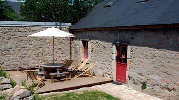 Een stenen gebouw met een rode deur, een houten terras met tuinmeubilair en een grote parasol.