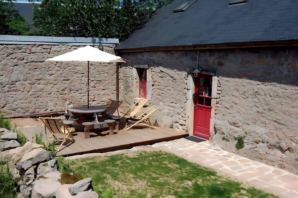 A stone building with a red door, a wooden deck with outdoor furniture, and a large umbrella. - Morvan Natural Park Gîte with Private Terrace, Garden, and Wi-Fi (Quarré-les-Tombes)