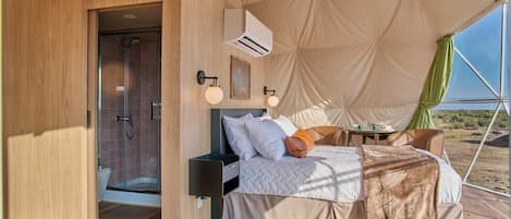 A tented accommodation with a bed, bedside table, lamp, and a view of the desert.