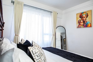 A bedroom with a bed, a large mirror, a painting on the wall, and a window with curtains. - Modern Kilimani Apt with Stunning Nairobi Views! Guest Friendly! (Nairobi)