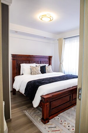 A hotel room with a wooden bed, white bedding, a patterned rug, and a ceiling light. - Modern Kilimani Apt with Stunning Nairobi Views! Guest Friendly! (Nairobi)