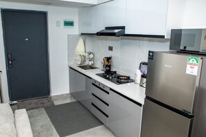Una cocina compacta con un refrigerador de acero inoxidable, una estufa a gas y un microondas. Hay un lavamanos pequeño con una toalla colgando encima. La cocina tiene armarios blancos y una puerta oscura.