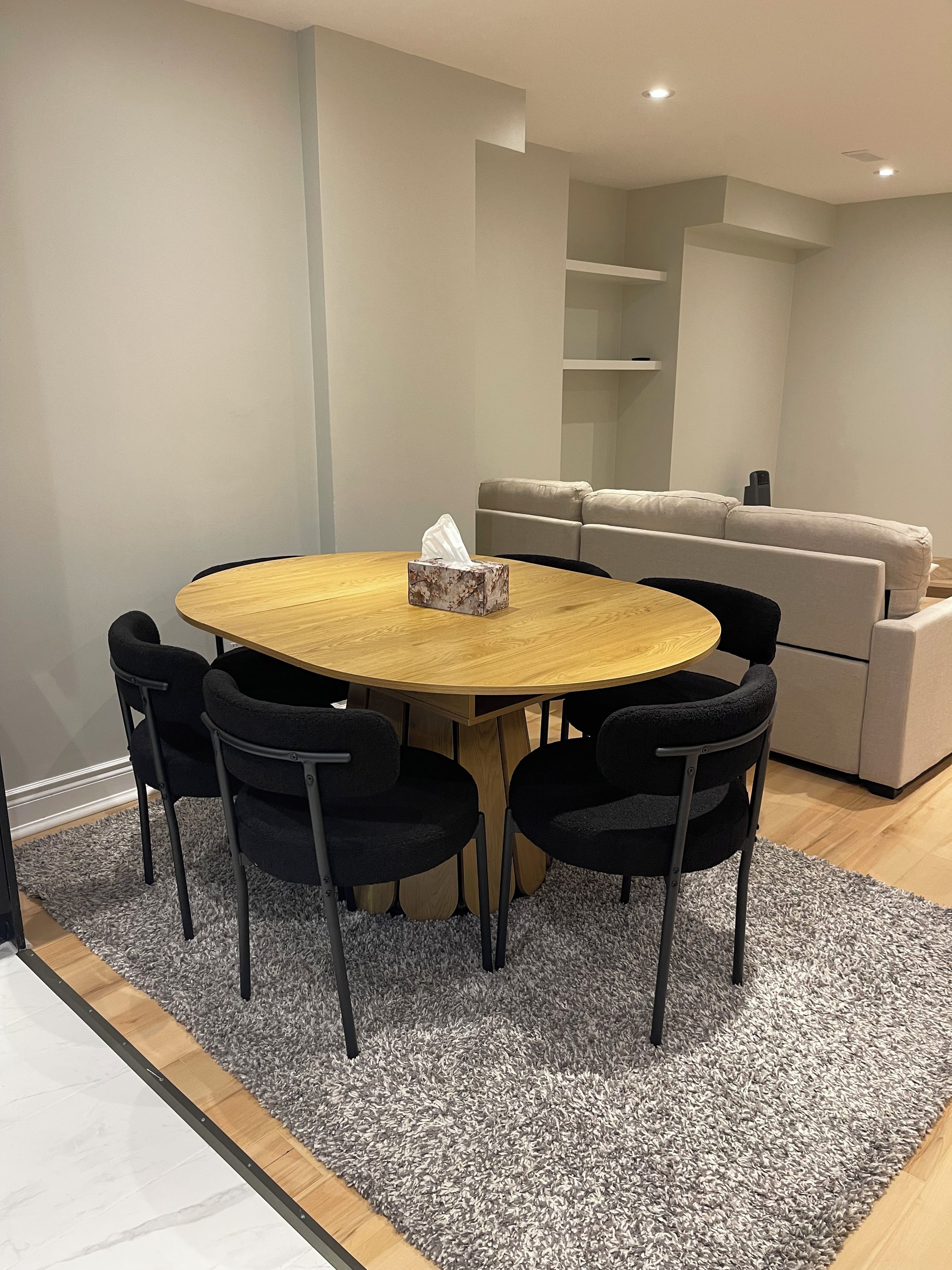 A modern dining area with a round wooden table, four black chairs, and a grey textured rug.