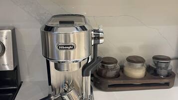 A DeLonghi espresso machine on a kitchen counter with a marble backsplash and a wooden tray with jars.