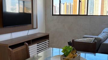 Uma sala de estar moderna com uma mesa de jantar de vidro, uma televisão de tela plana fixada na parede, um sofá e uma grande janela com vista para a cidade.