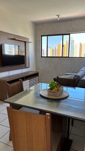 Uma sala de estar moderna com uma mesa de jantar de vidro, uma televisão de tela plana fixada na parede, um sofá e uma grande janela com vista para a cidade.