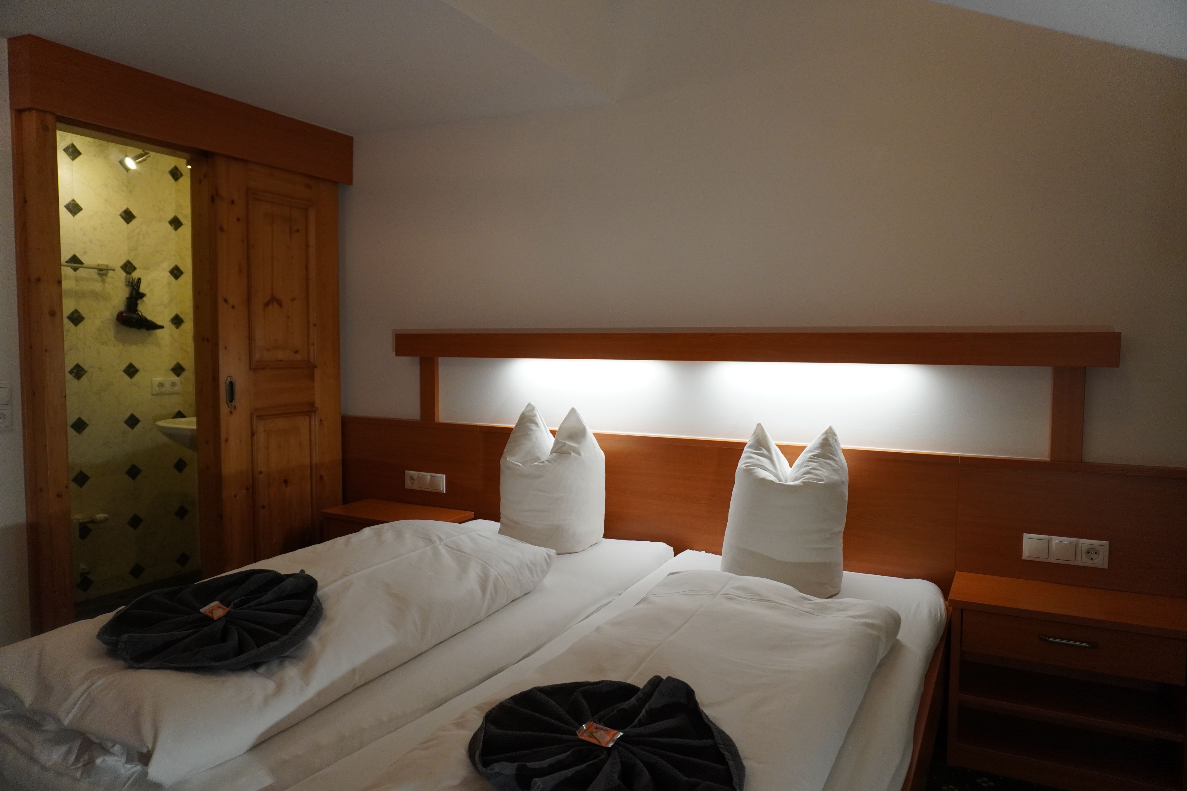 A hotel room with two beds, a wooden headboard, a nightstand, a bedside table, and a bathroom with a wooden door.