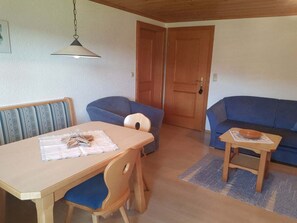 A room with a wooden dining table, two blue sofas, a small wooden table with a bowl, and a closed wooden door. - Holiday Apartment With 1 Bedroom (Drachselsried)