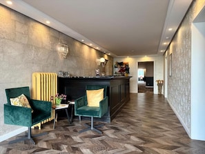 Un hall d’hôtel avec un comptoir d’accueil, deux fauteuils verts, une petite table sur laquelle est posé un vase de fleurs et un sol à motifs en chevrons.
