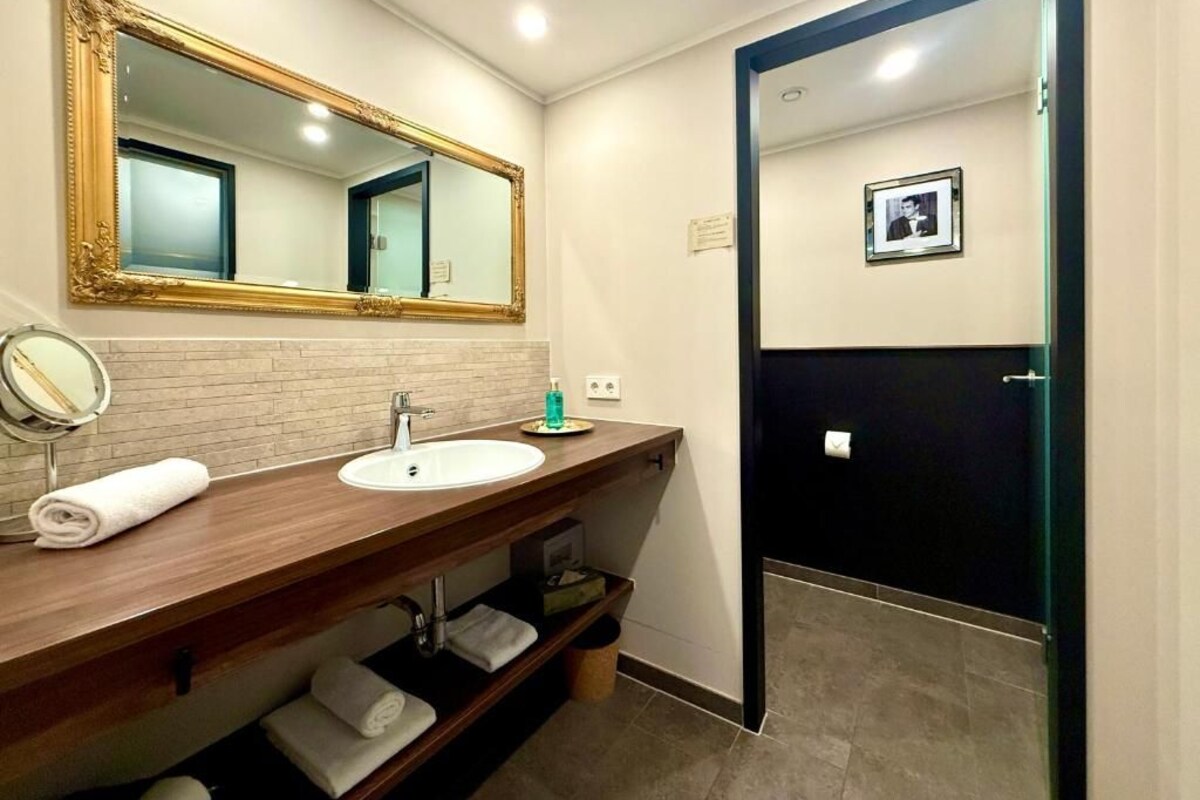 Een badkamer met een houten wastafelmeubel, een grote spiegel, een wastafel en een zwarte deur naar het toilet.
