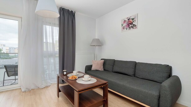 A modern living room with a grey sofa, a wooden coffee table, and a view of the city through large windows. - Łódź | Kazimierza Odnowiciela 13 | Family Apartment | Balcony (Łódź)
