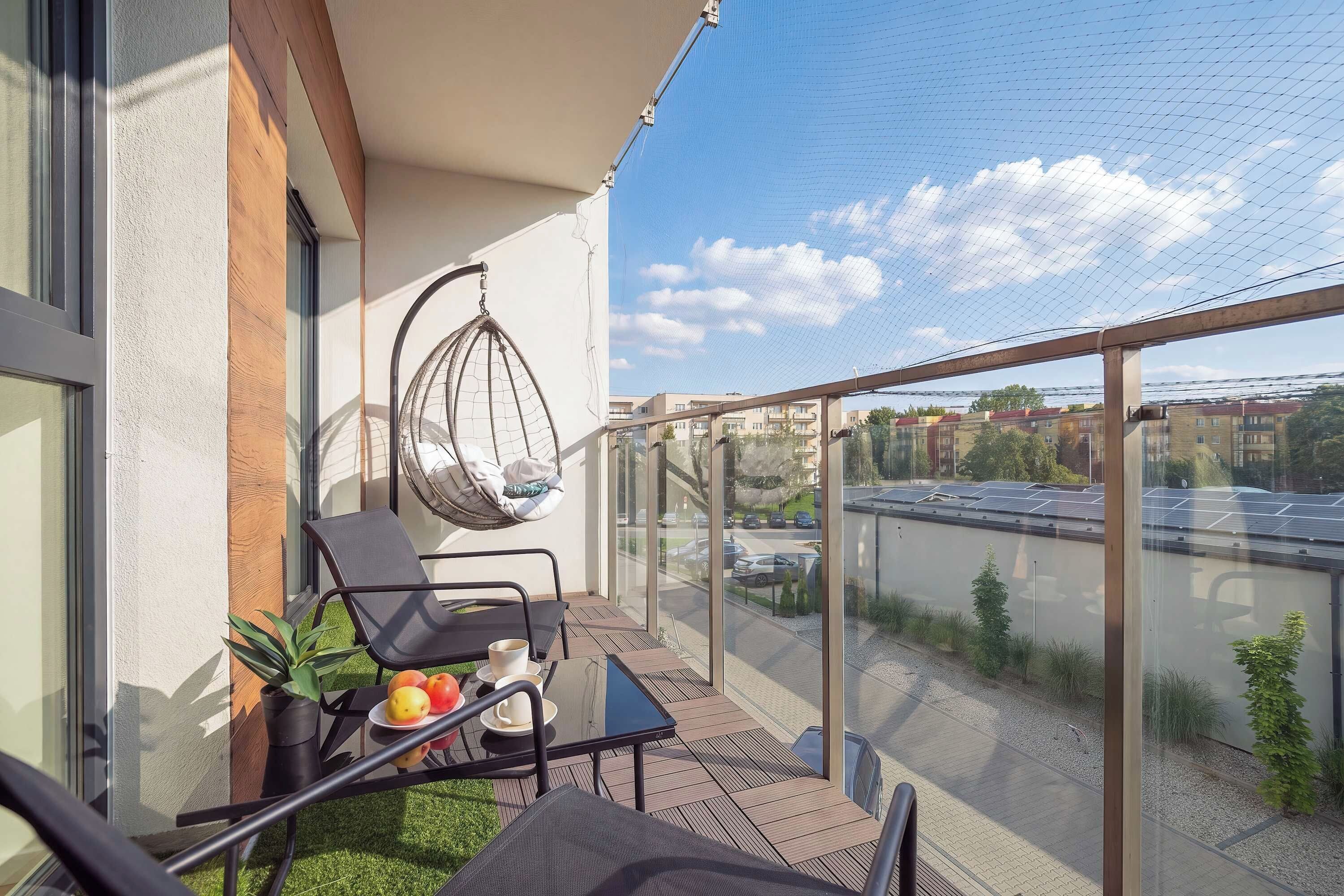 A balcony with a glass railing, a hanging chair, a table with a chair, and a potted plant.