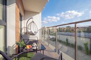 A balcony with a glass railing, a hanging chair, a table with a chair, and a potted plant.