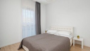 A minimalist bedroom with a large bed, a bedside table, and a window with curtains. - Łódź | Kazimierza Odnowiciela 13 | Family Apartment | Balcony (Łódź)