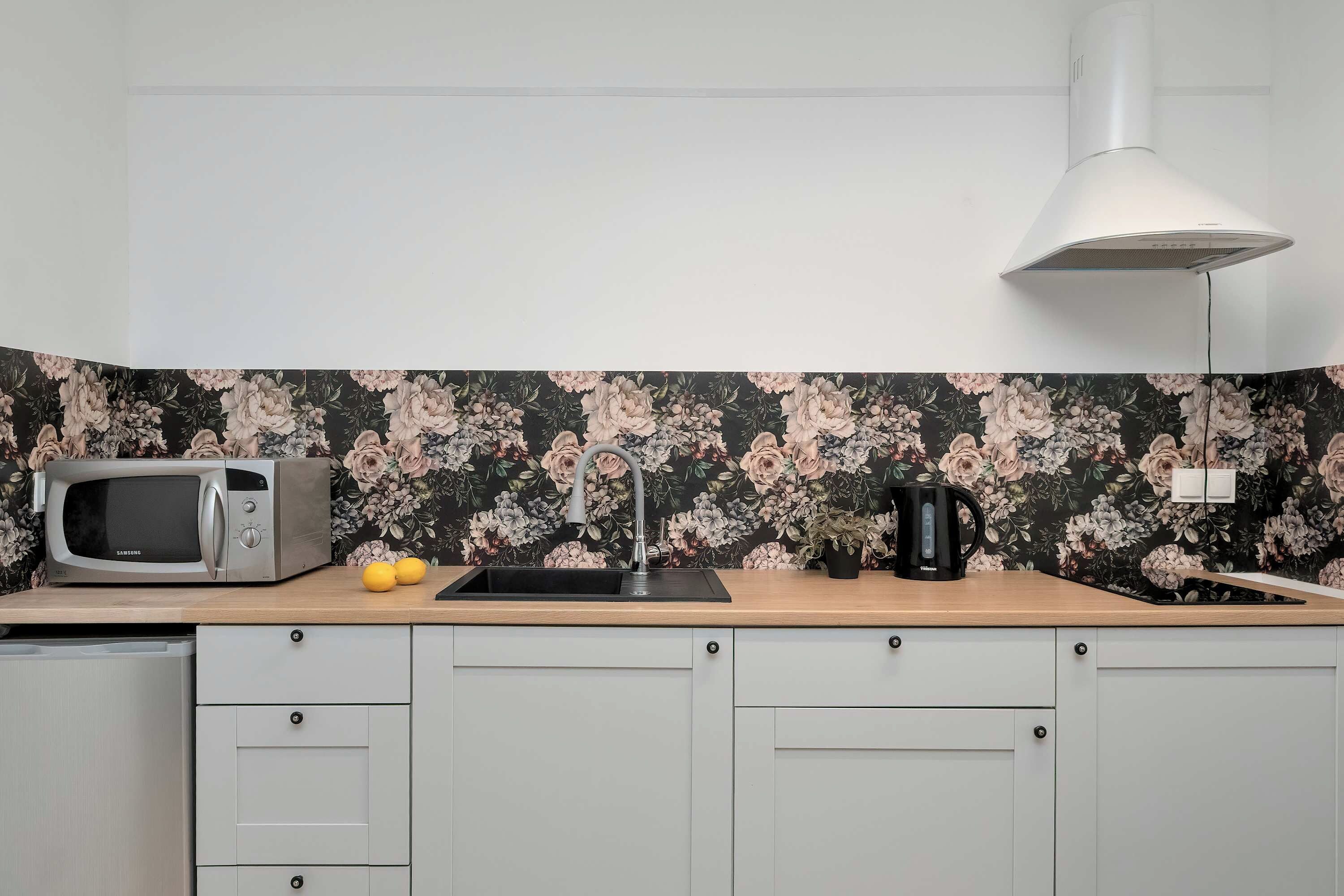Een keuken met een magnetron, spoelbak en waterkoker op het aanrecht, met een bloemenachtergrond en lichte kastjes.