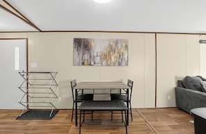 A room with a dining table and chairs, a grey sofa, a wall-mounted abstract painting, and a metal drying rack. - 
Fully Furnished 3BR/2BA Monthly Workforce Housing Near Holly Ridge Data Center (Monroe)