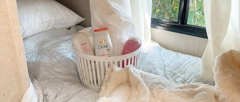 A bed with a fluffy white blanket, a white basket with toiletries, and a window with curtains.