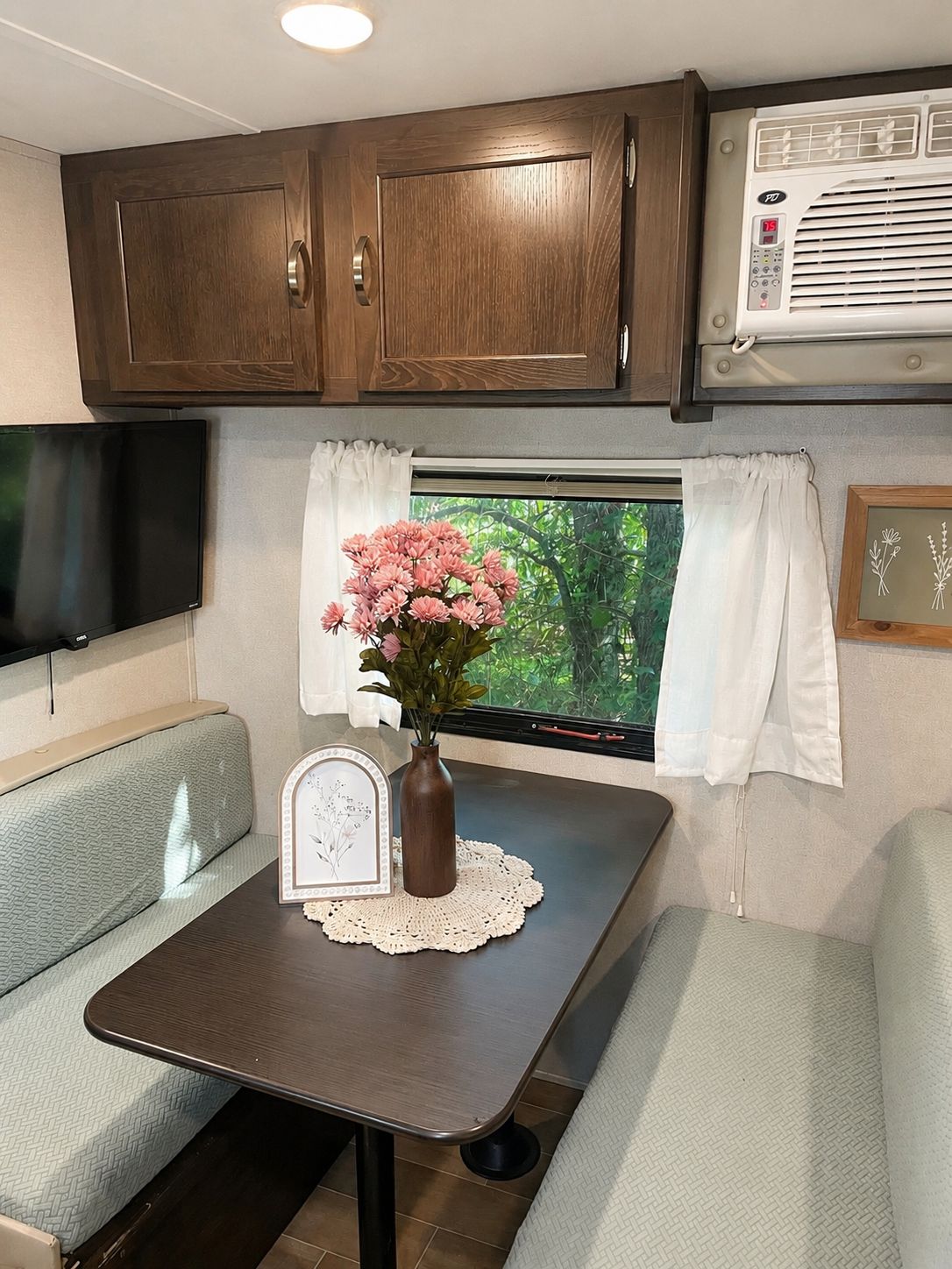 A compact RV interior with a dining table, a small TV, and a window with curtains.