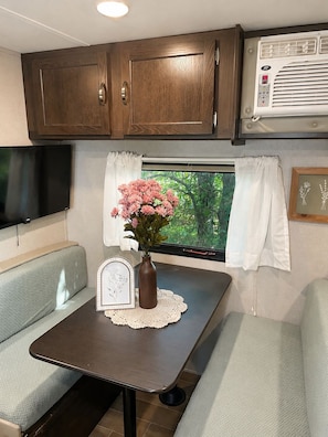 Interior compacto de una caravana con una mesa de comedor, un televisor pequeño y una ventana con cortinas.
