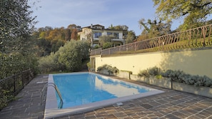 Una piscina rectangular con una escalera de metal, rodeada por un camino de piedra y una valla de metal.