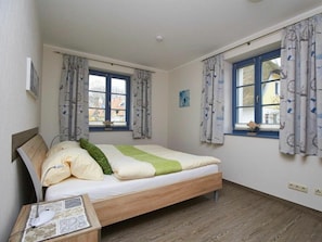 A bedroom with a bed, two windows with curtains, a bedside table with a lamp, and a small picture frame. - New Row 43 Villa Regulus Ground Floor (Zingst)