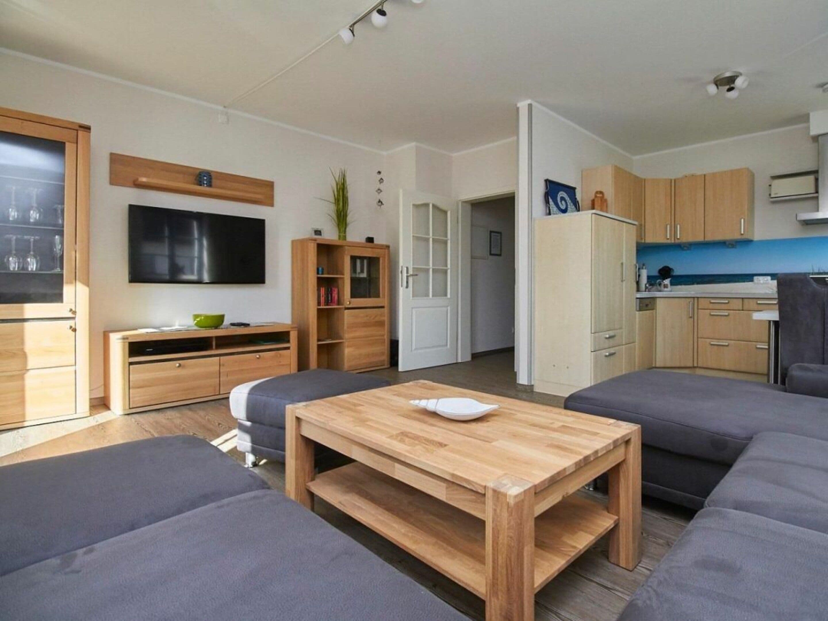 A modern living room with a flat-screen TV, wooden furniture, and a kitchen area with cabinets and a refrigerator.