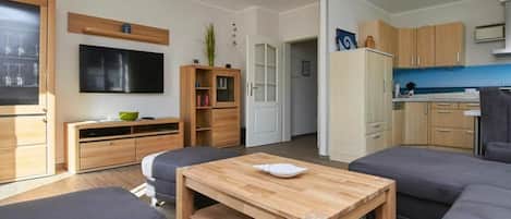 A modern living room with a flat-screen TV, wooden furniture, and a kitchen area with cabinets and a refrigerator.