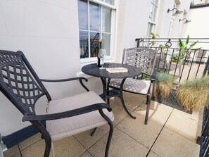 En terrasse med et rundt bord og to stole, et vindue med udsigt til en bygning, og et lille område med planter.