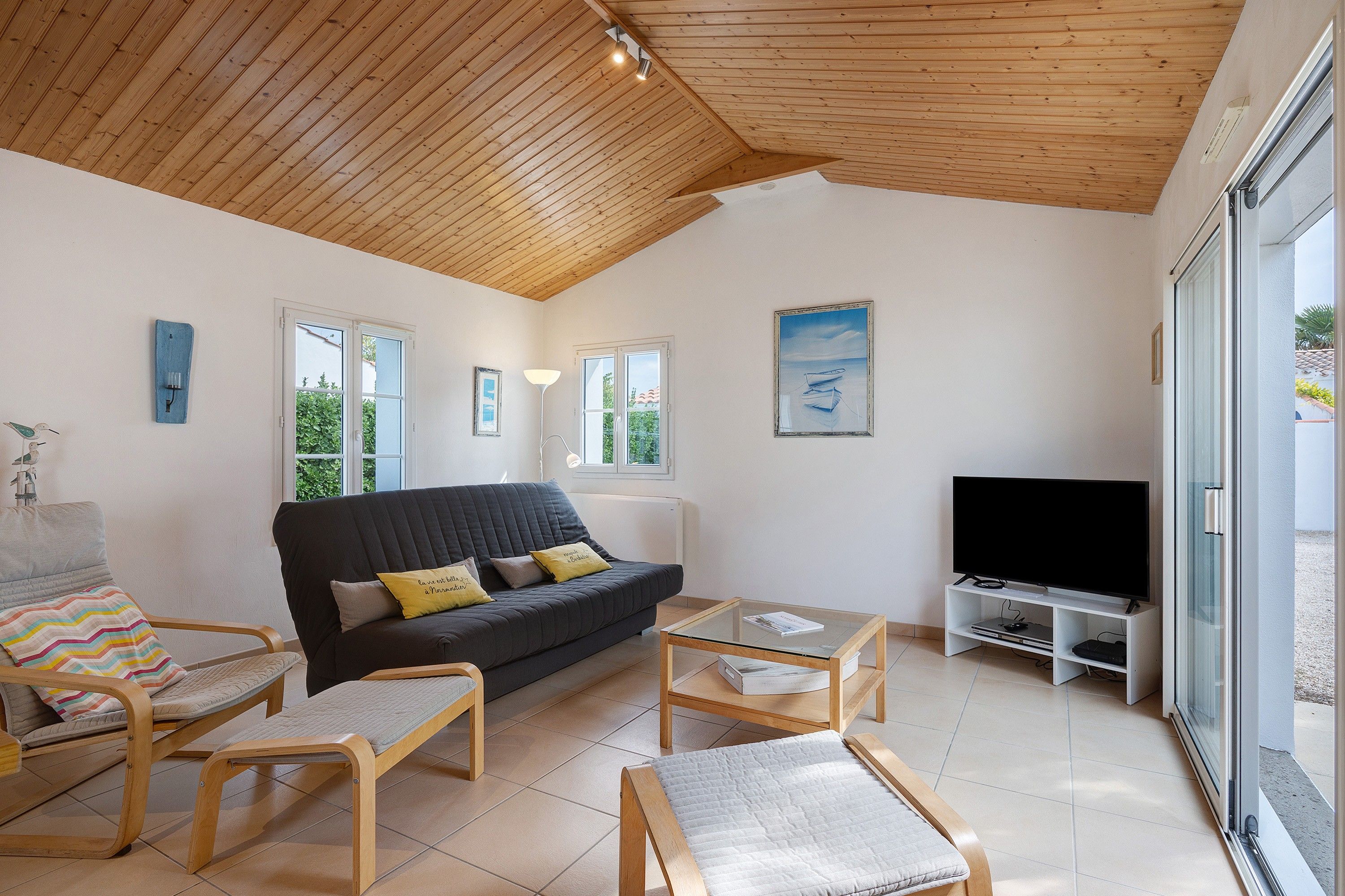 Ett vardagsrum med ett träpanelat tak, en soffa, två fåtöljer, ett soffbord, en tv och ett fönster med utsikt över grönområden.