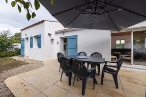 Una veranda esterna con tavolo e sedie, un ombrellone grande e un edificio bianco con persiane blu.