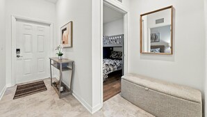 Ein Flur mit einer weißen Tür, einem Konsolentisch mit einer Pflanze, einer Bank und einem Bett mit gemustertem Bettzeug im angrenzenden Zimmer.