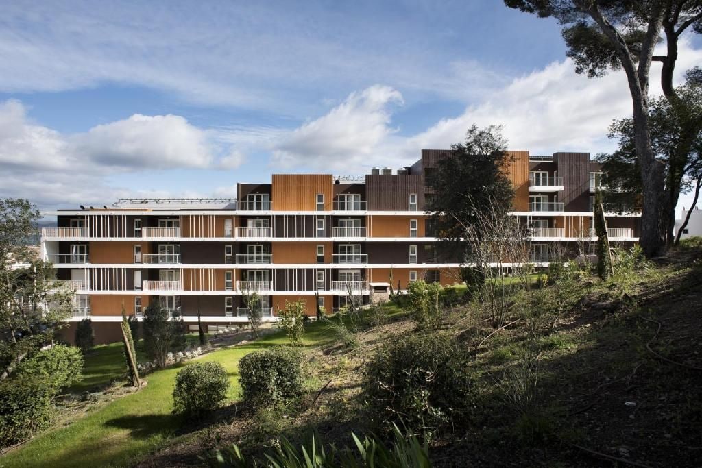 Un edificio residencial de varios pisos con balcones, rodeado de follaje y árboles.