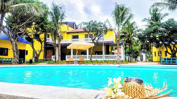 Ein Swimmingpool mit Liegestühlen und Sonnenschirmen, umgeben von Palmen und einem gelben Gebäude mit weißem Zaun.