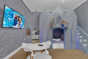 A room with a television displaying animated characters, a white table with chairs, and a bed with a decorative headboard. - Themed 8BR Home w/ Private Pool & Custom Game Room (Kissimmee)