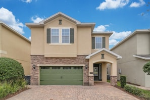 A two-story residential building with a green garage door, stone accents, and a clear blue sky. - Themed 8BR Home w/ Private Pool & Custom Game Room (Kissimmee)