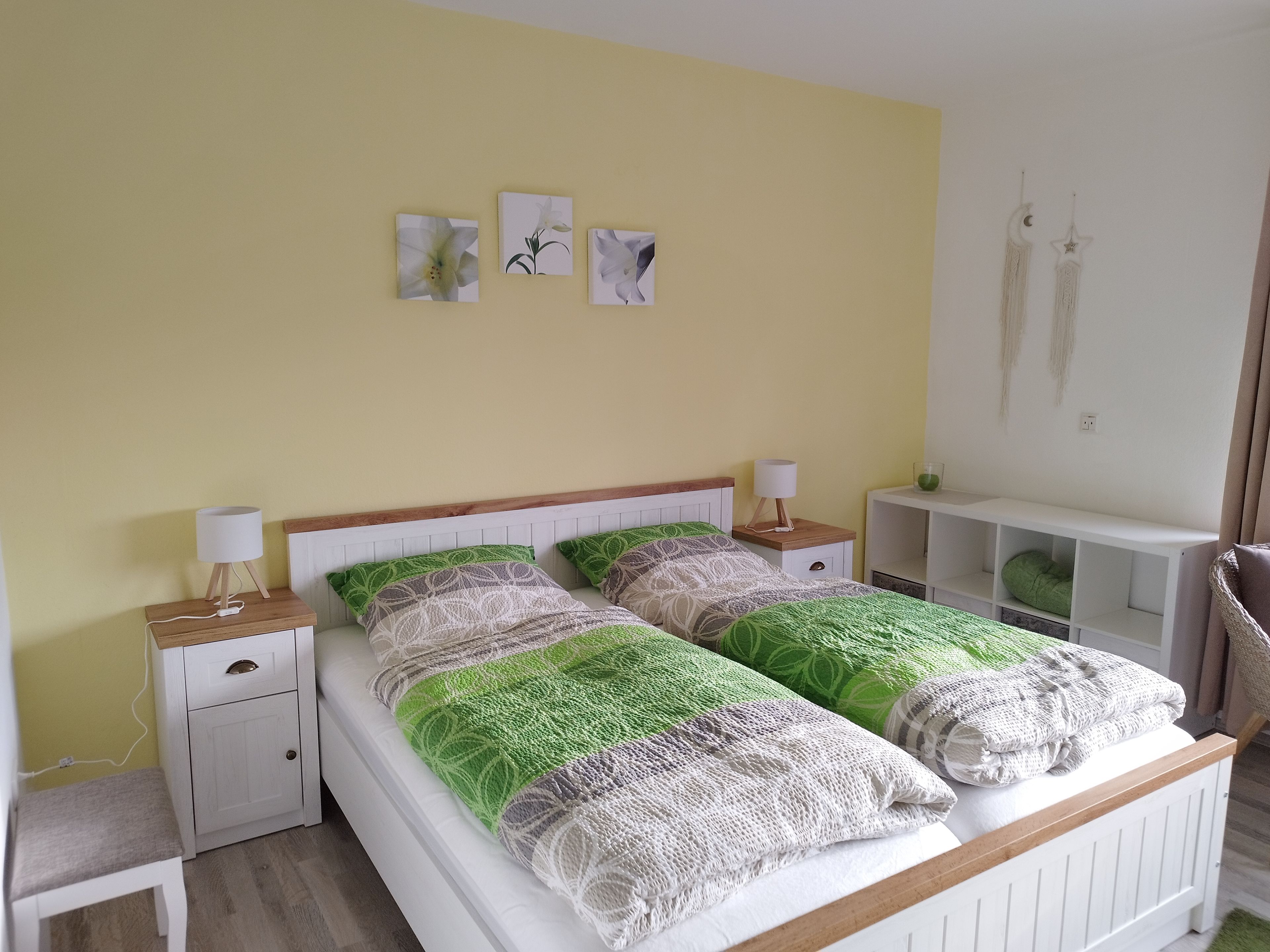 Een tweepersoonsbed met een houten hoofdbord, twee nachtkastjes met lampen, een witte bijzettafel, een groene kussen en een witte plankunit.