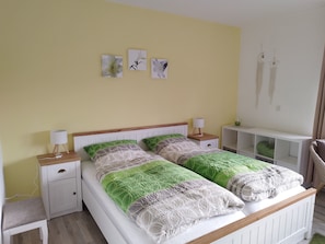 A double bed with a wooden headboard, two bedside tables with lamps, a white nightstand, a green cushion, and a white shelf unit.