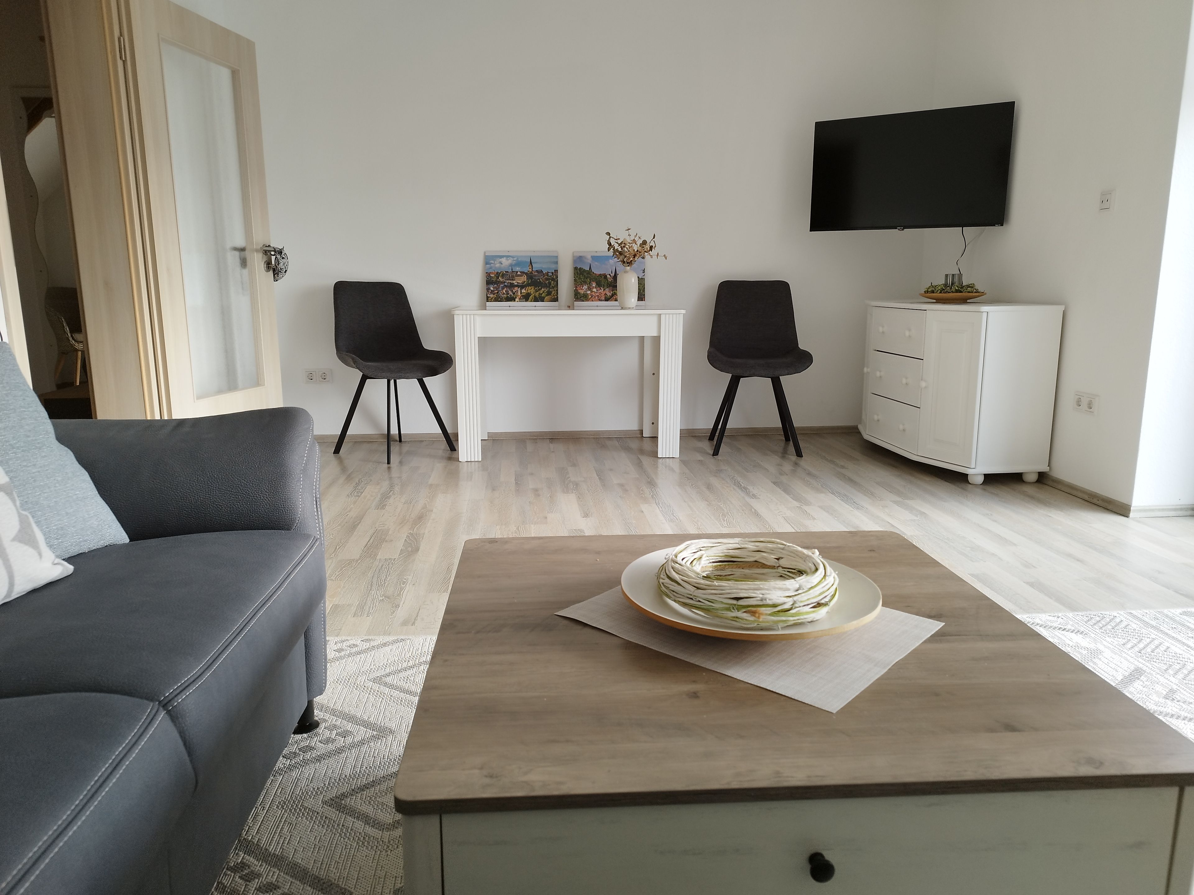 A modern living room with a grey sofa, a wooden coffee table with a decorative item, a white desk with two chairs, a TV mounted on the wall, and a white cabinet.