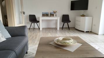 A modern living room with a grey sofa, a wooden coffee table with a decorative item, a white desk with two chairs, a TV mounted on the wall, and a white cabinet.