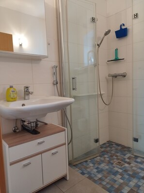 Ein Badezimmer mit einer gläsernen Duschkabine, einem weißen Waschbecken mit Holzablage und einem gefliesten Boden.
