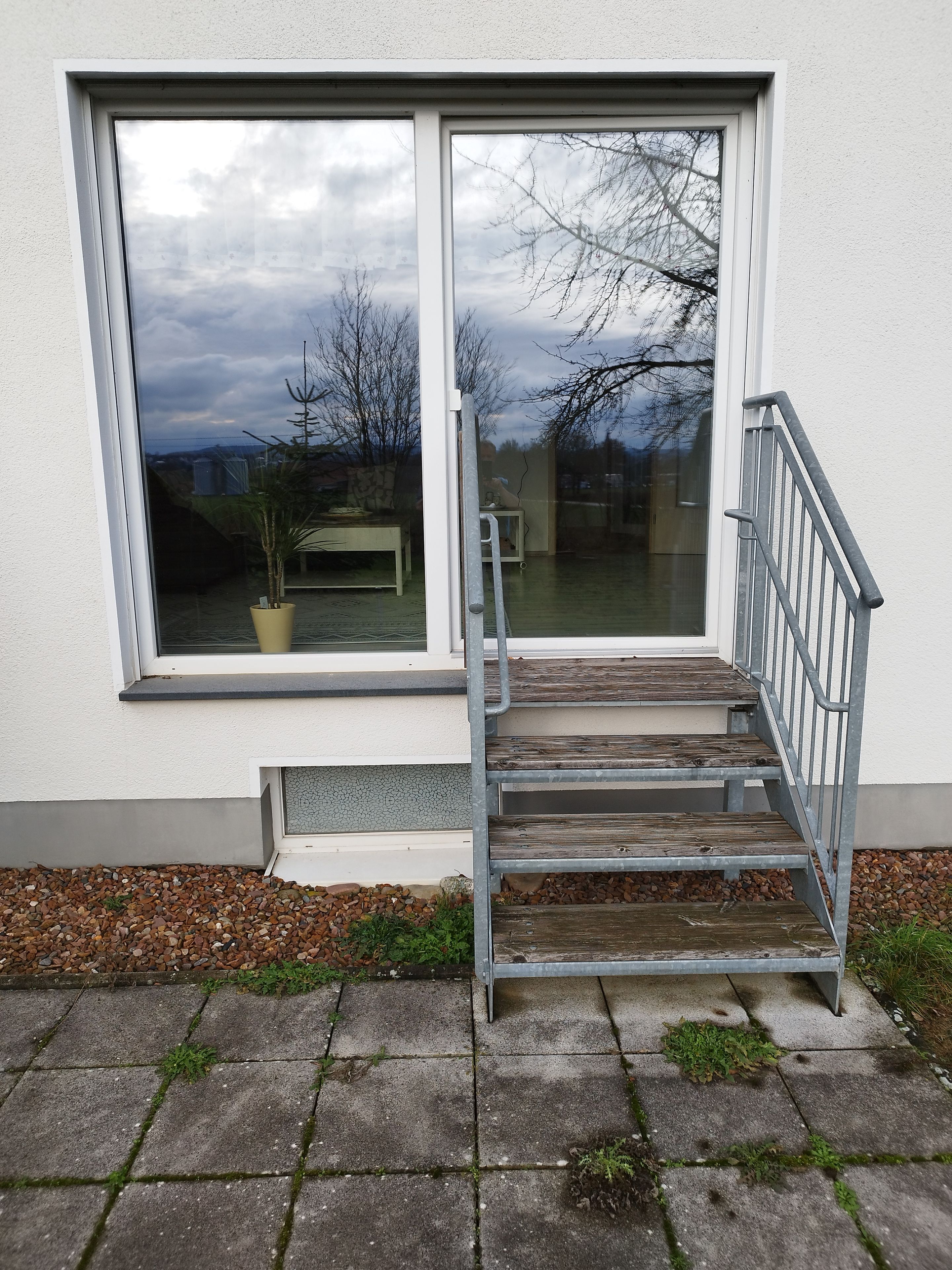 Eine Treppe führt zu einem Fenster mit weißem Rahmen und Blick auf Bäume und einen bewölkten Himmel.