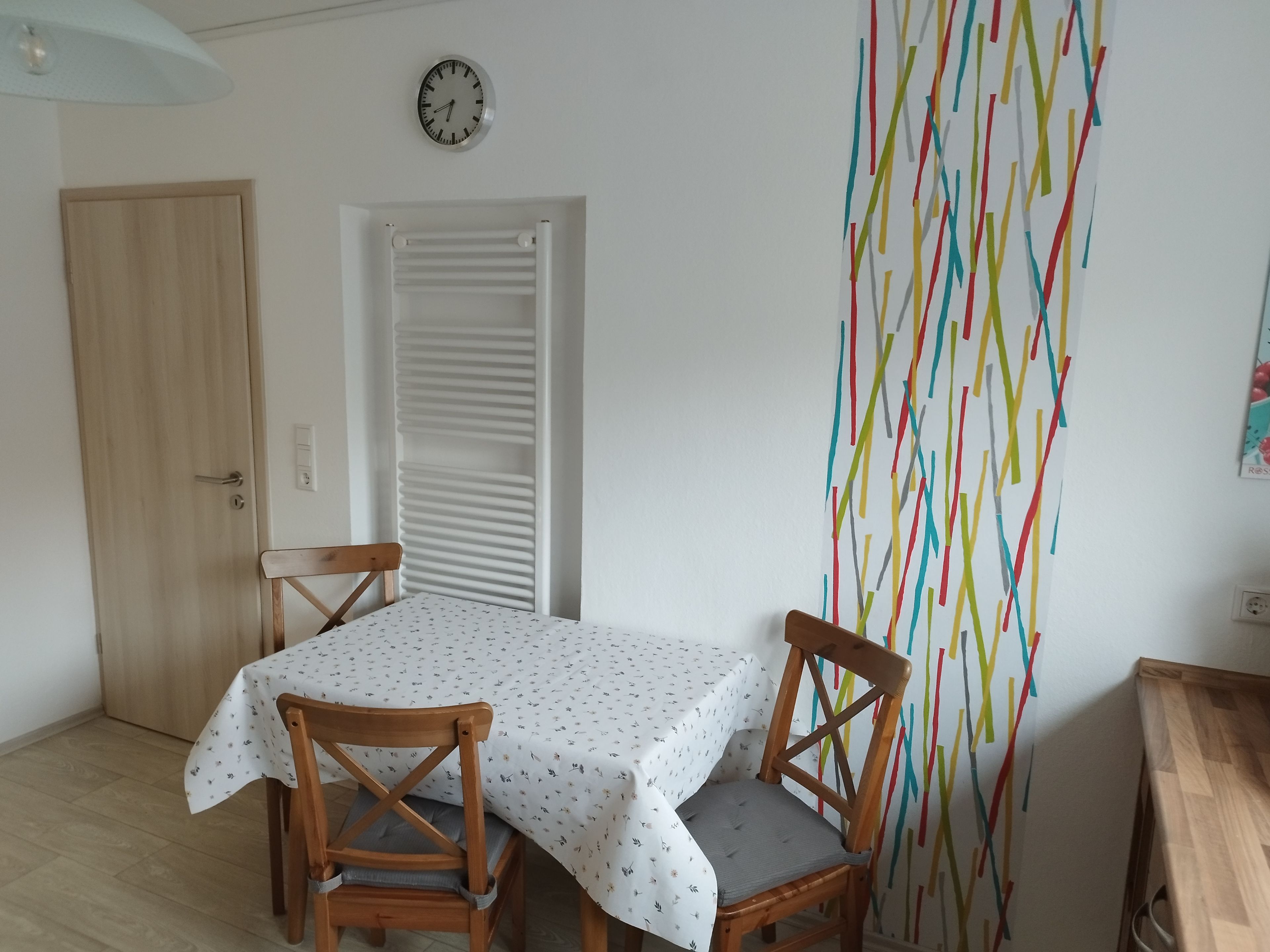 Een eetgedeelte met een tafel voor twee, een klok aan de muur en een kleurrijke abstracte wanddecoratie.