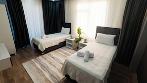 Habitación de hotel con dos camas individuales, una mesita de noche con lámpara, un ventanal con cortinas y suelo de madera.