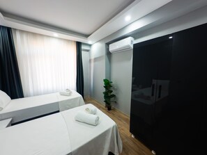 A hotel room with two beds, a large window with curtains, an air conditioning unit, a plant, and a black wardrobe.