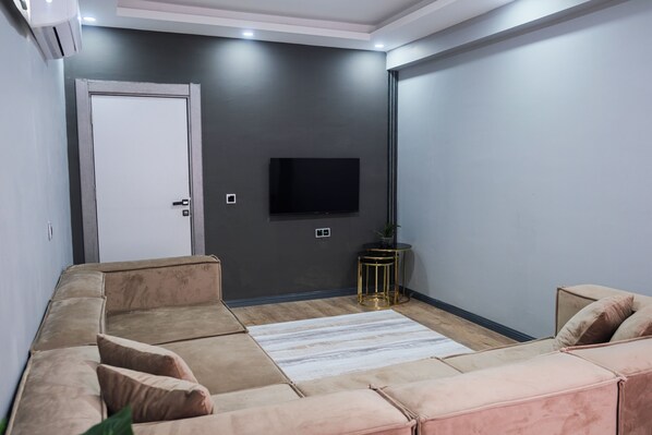 A modern living room with a sectional sofa, a flat-screen TV mounted on the wall, and a small round table with a plant.