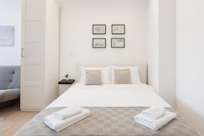 Quarto de hotel com cama, mesa de cabeceira com planta e sofá cinza. Há quatro quadros na parede.