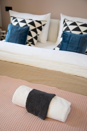 Cama bem arrumada com travesseiro estampado em branco e preto, um travesseiro azul e uma toalha dobrada sobre um espalha cama rosa texturizado.