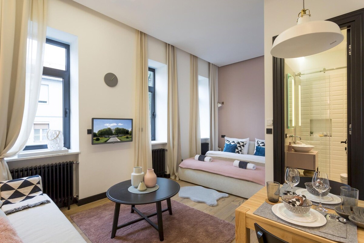 A well-lit hotel room with a bed, bedside table, and a dining area with a table set for two.
