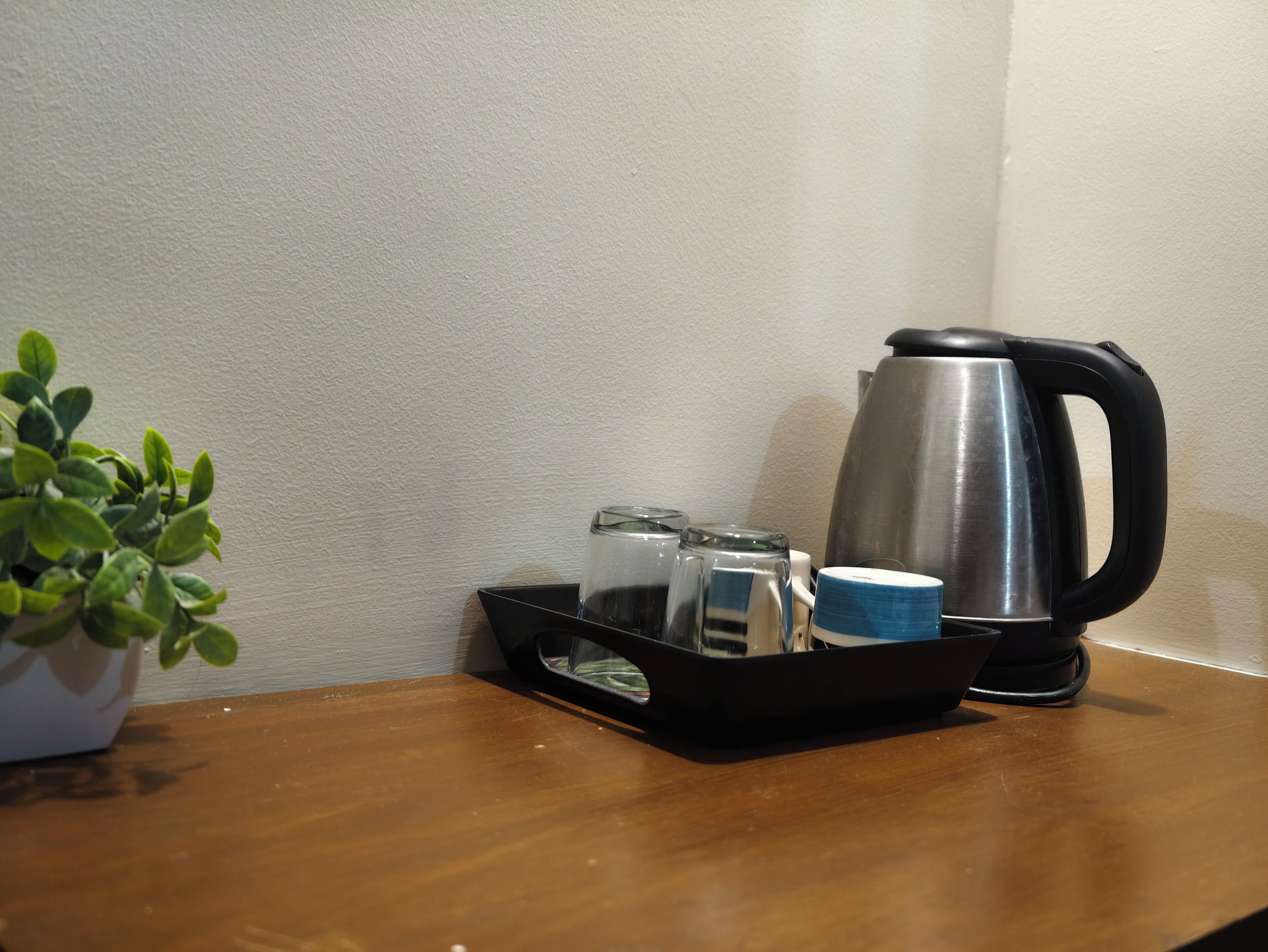 Una mesa de madera con una bandeja negra que sostiene un hervidor de acero inoxidable, un tarro de vidrio y una taza azul. Se ve una planta en maceta a la izquierda.