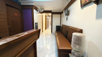 Zona de recepción de un hotel con mostrador de madera, un banco, dispensador de agua y un televisor en la pared.